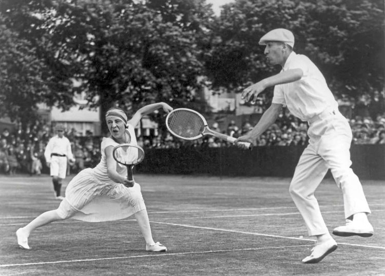 Eine Dame und ein Herr in altertümlicher Tenniskleidung bei einem Spiel.
