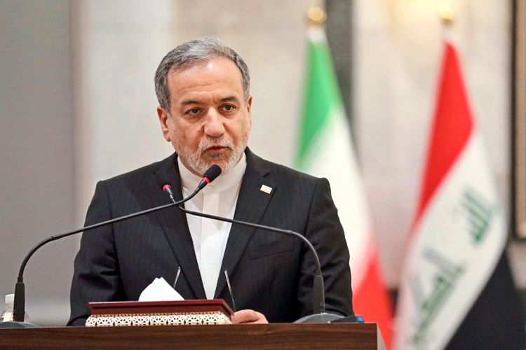 Ein Mann in einem dunklen Anzug spricht bei einer Pressekonferenz hinter einem Rednerpult. Im Hintergrund sind unscharf die iranische und die irakische Flagge zu sehen.
