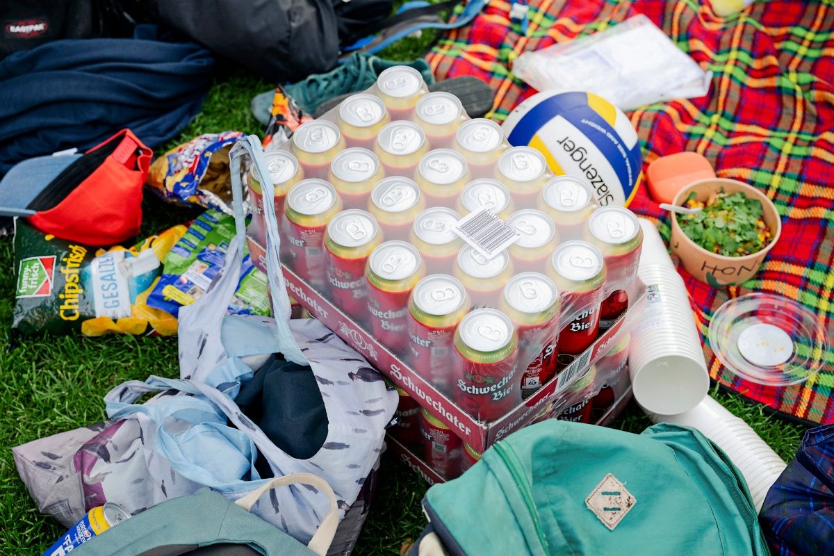 Ein Picknick-Setup auf einer Wiese. Zu sehen sind eine Palette mit Bierdosen, ein Volleyball, eine Schüssel mit Salat, Plastikbecher, verschiedene Chipspackungen und Taschen auf einer rot-karierten Decke.