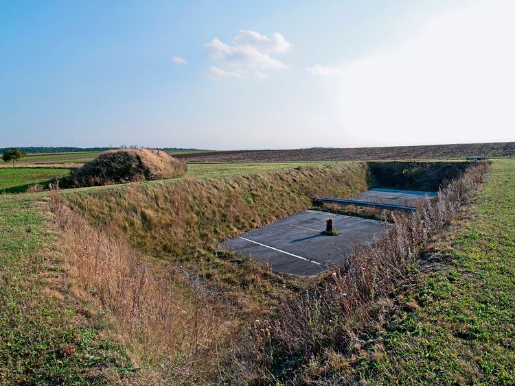 Eine verlassene, eingesenkte Fläche im Freien, die wie ein alter Parkplatz aussieht, umgeben von grasbewachsenen Böschungen und Wildpflanzen. Im Hintergrund sind ein offenes Feld und ein Baum erkennbar, mit einem klaren Himmel darüber.
