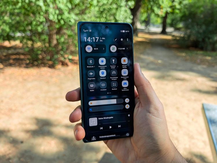 Eine Hand hält ein Smartphone in einem Park. Der Bildschirm zeigt das Kontrollzentrum des Geräts mit verschiedenen Einstellungen wie WLAN, Bluetooth und Taschenlampe. Bäume und ein Weg im Hintergrund sind unscharf zu erkennen.