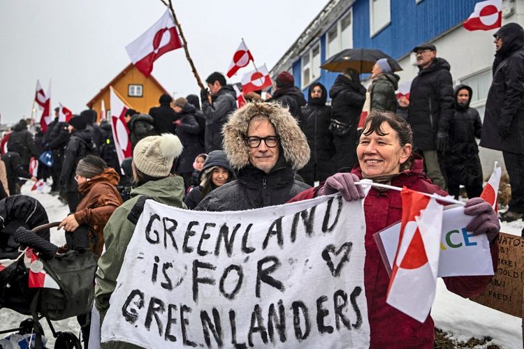 Menschen nehmen an einem Protest in Nuuk, Grönland, teil. Im Vordergrund halten Teilnehmer ein Banner mit der Aufschrift „Greenland is for Greenlanders“ und tragen Kleidung, die auf kaltes Wetter hinweist. Im Hintergrund sind weitere Protestierende mit grönländischen Flaggen erkennbar. Schneebedeckter Boden und Gebäude rahmen die Szene ein.