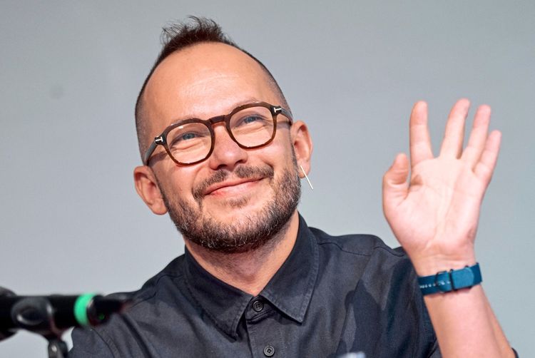 Ein Mann mit Brille und kurzem Bart sitzt lächelnd vor einem Mikrofon und winkt. Er trägt ein dunkles Hemd und eine blaue Uhr. Hintergrund ist neutral gehalten.