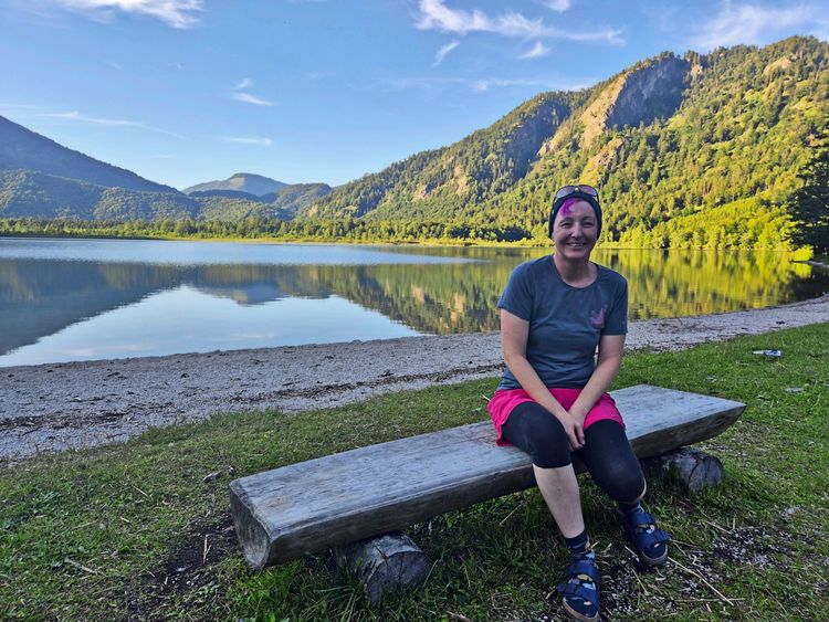 Eine Frau sitzt auf einer Holzbank am Ufer des Offensees, umgeben von grünen Bergen und ruhigem, spiegelndem Wasser. Der Himmel ist klar und blau. Die Umgebung strahlt eine ruhige, natürliche Atmosphäre aus.