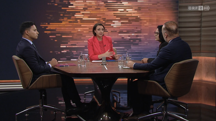 Eine Diskussionsrunde im TV-Studio mit vier Personen, moderiert von einer Frau in rotem Blazer. Drei Gäste, zwei Männer und eine Frau, sitzen an einem runden Tisch. Im Hintergrund ist eine abstrakte, beleuchtete Wand zu sehen.