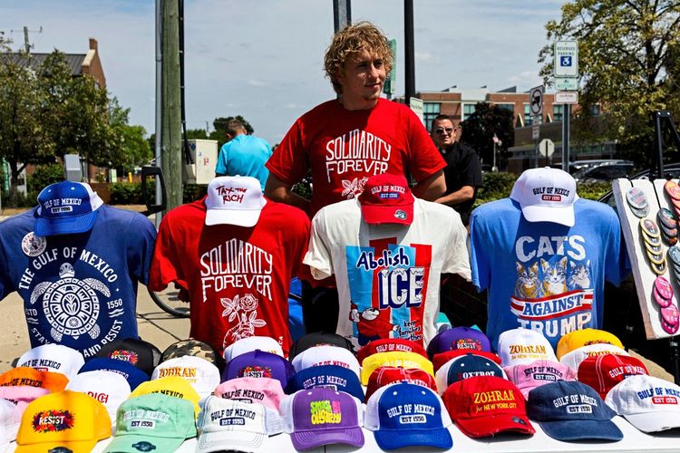 Ein Verkaufsstand mit politischen T-Shirts, Mützen und Buttons vor einer Wahlkampfveranstaltung in Ferndale, Michigan, am 25. Juli 2026. Die Artikel haben Aufschriften wie „Solidarity Forever“, „Abolish ICE“ und „Cats Against Trump“. Ein Mann in einem roten T-Shirt steht hinter dem Stand.