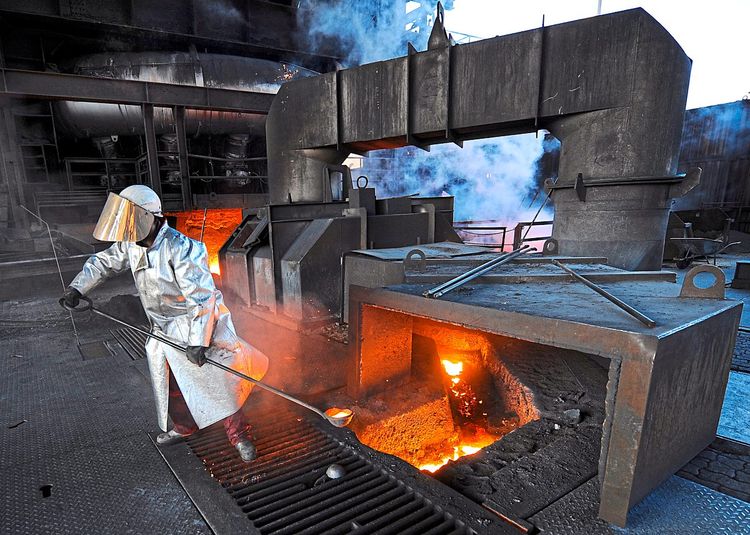Ein Stahlarbeiter in Schutzkleidung hat eine Kelle mit flüssigem, gleißendem Metall in den Händen.