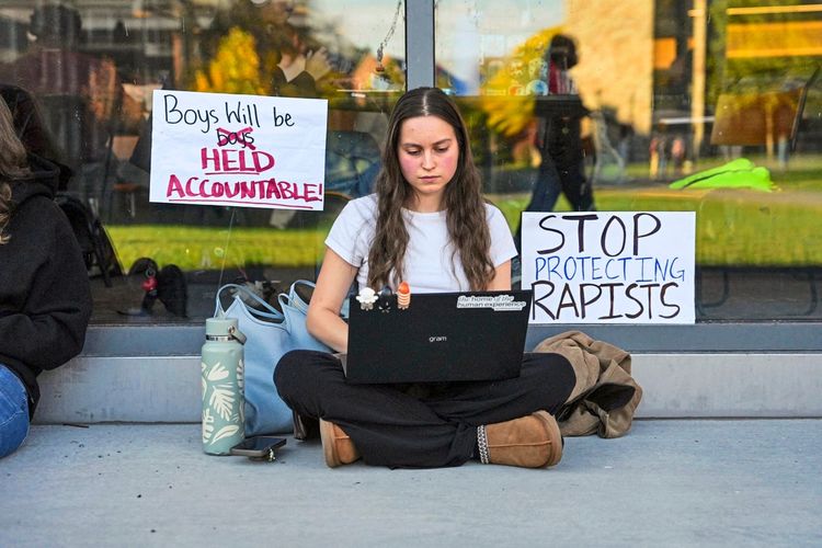 Eine Studentin der Cornell University sitzt auf dem Boden vor einer Glasfassade, arbeitet an einem Laptop und ist von Protestplakaten umgeben. Auf den Plakaten stehen Botschaften wie „Boys will be held accountable!“ und „Stop protecting rapists“. Daneben steht eine Wasserflasche und eine Tasche. Die Szene findet im Rahmen eines Campus-Protests gegen den Umgang der Universität mit einem sexuellen Übergriff statt.