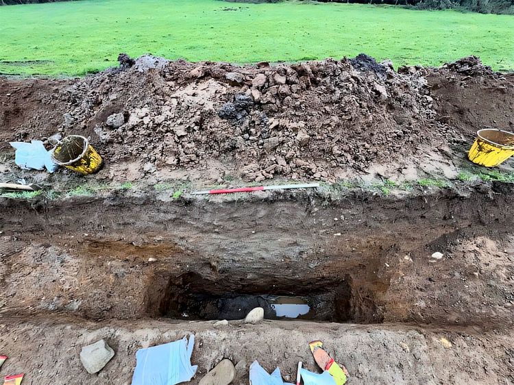 Bei den Ausgrabungen ist ein freigelegter, länglicher Graben zu sehen, der teilweise mit Wasser gefüllt ist. Erde ist aufgeschüttet, im Hintergrund ist eine grasbewachsene Fläche zu erkennen. Gelbe Eimer und Werkzeuge liegen um die Grabung verteilt.