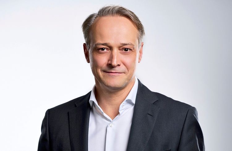 Ein Mann mittleren Alters trägt ein weißes Hemd und ein dunkelblaues Sakko. Er hat kurzes, glattes, grau-blondes Haar und schaut mit einem dezenten Lächeln direkt in die Kamera. Der Hintergrund ist einfarbig grau.
