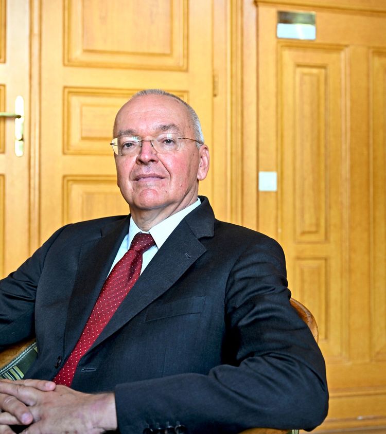 Ein Mann in formeller Kleidung, mit Anzug und roter Krawatte, sitzt in einem Stuhl vor einer Wand mit hölzernen Türpanelen.