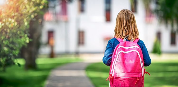 Ein Mädchen mit einem Schulrucksack steht mit dem Rücken zum Betrachtenden vor einem Schulgebäude