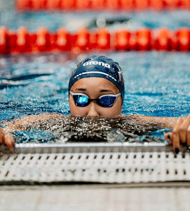 Eine Schwimmerin mit Schwimmhaube und Schwimmbrille hält sich am Beckenrand eines Schwimmbads fest. Im Hintergrund sind rote Schwimmbahnenmarkierungen sichtbar.