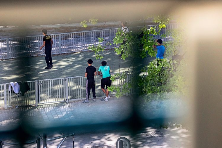 Eine Gruppe von jungen Menschen befindet sich in einem eingezäunten Außenbereich, umgeben von Bäumen. Einige stehen oder lehnen am Zaun, während eine Person im Hintergrund auf dem Boden des Bereichs geht. Die Szene scheint Teil eines Programms oder einer Aktivität im Jugendstrafvollzug zu sein.