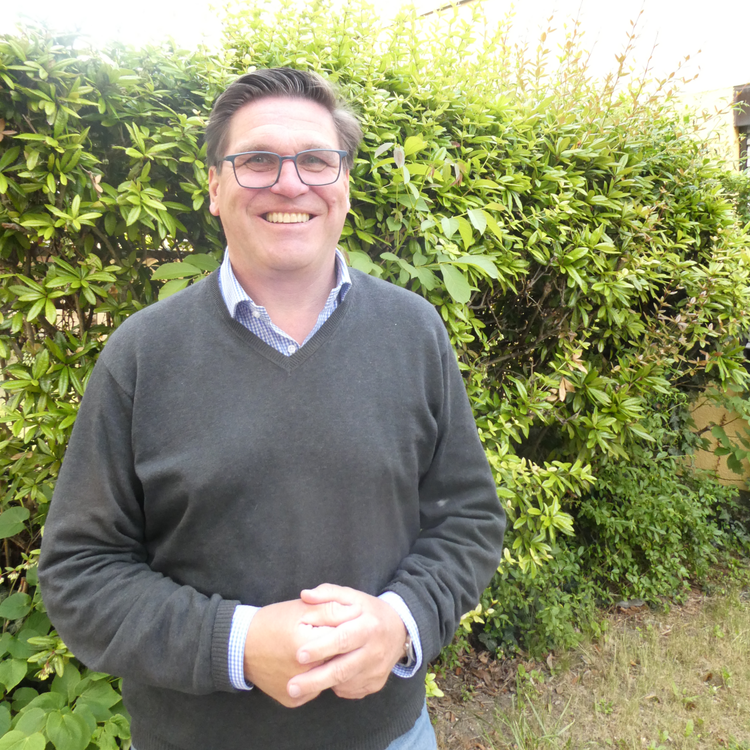 Ein lächelnder Mann mit Brille steht in einem Garten vor einer grünen Hecke. Er trägt einen dunkelgrauen Pullover über einem karierten Hemd.