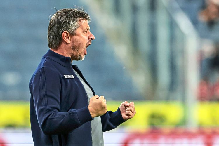 Ein Fußballtrainer in blauer Kleidung jubelt mit geballten Fäusten auf einem Spielfeld während eines Spiels. Im Hintergrund sind verschwommene Zuschauerränge zu sehen.