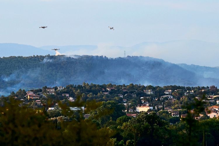 Rauch steigt von einem Waldbrand in der Nähe von Arc-sur-Argens, Südfrankreich, auf. Drei Löschflugzeuge sind im Einsatz und werfen Löschmittel über das betroffene Gebiet ab. Im Vordergrund ist eine Siedlung mit Häusern und Bäumen zu sehen, dahinter bewaldete Hügel.