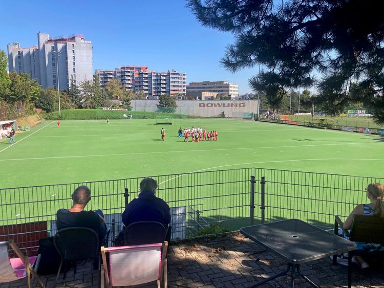 Ein Hockeyfeld bei sonnigem Wetter, umgeben von städtischen Gebäuden und grünem Gelände. Zuschauer sitzen hinter einem Zaun auf Klappstühlen und blicken auf das Spielfeld, wo eine Gruppe von Spielern in Sportkleidung steht. Im Hintergrund ist eine Mauer mit der Aufschrift 