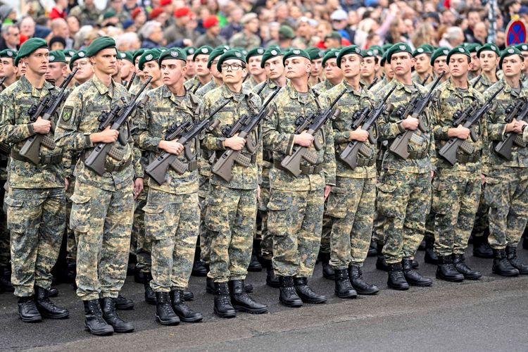 Eine Gruppe von Soldaten des österreichischen Bundesheeres in Tarnuniformen steht in Reihen, jeweils mit Gewehren ausgestattet. Im Hintergrund ist eine Menschenmenge zu sehen. Anlass ist eine Veranstaltung am Wiener Heldenplatz am Nationalfeiertag.