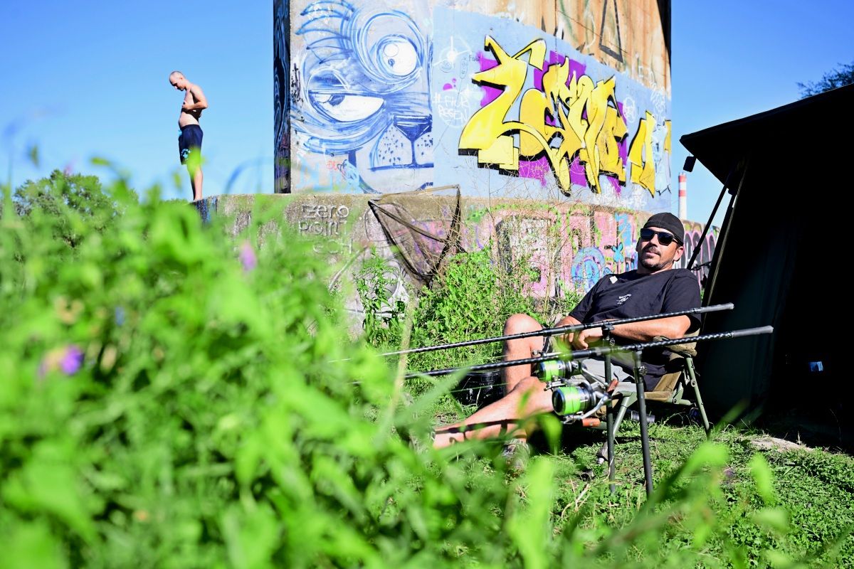 Ein Mann sitzt entspannt auf einem Angelstuhl mit Angelruten vor sich. Im Hintergrund ist eine Wand mit Graffiti und eine weitere Person in Badebekleidung zu sehen. Im Vordergrund befinden sich grüne Pflanzen.