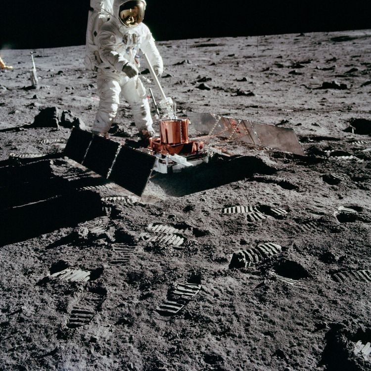 Ein Astronaut auf dem Mond steht vor einem zylinderförmigen Objekt, das mit Solarpaneelen versehen ist.