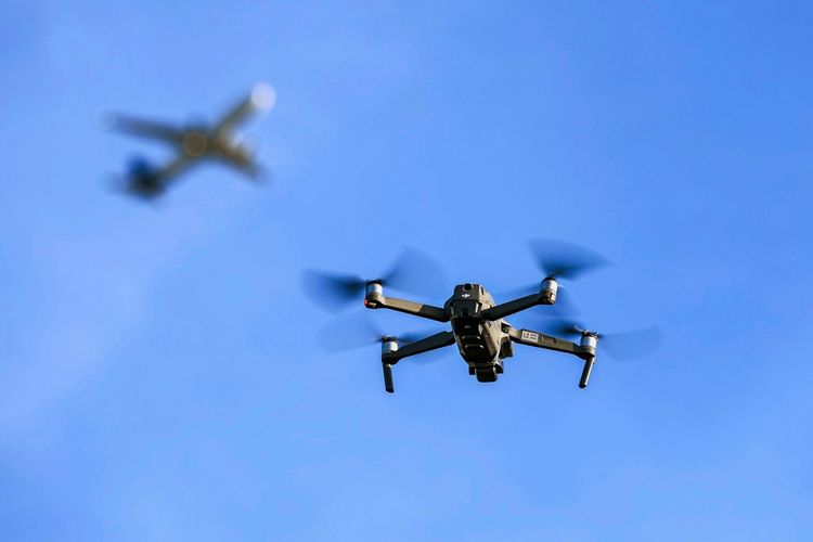 Eine Drohne fliegt vor einem unscharf im Hintergrund sichtbaren startenden Flugzeug nahe dem Flughafen Leipzig/Halle im blauen Himmel.