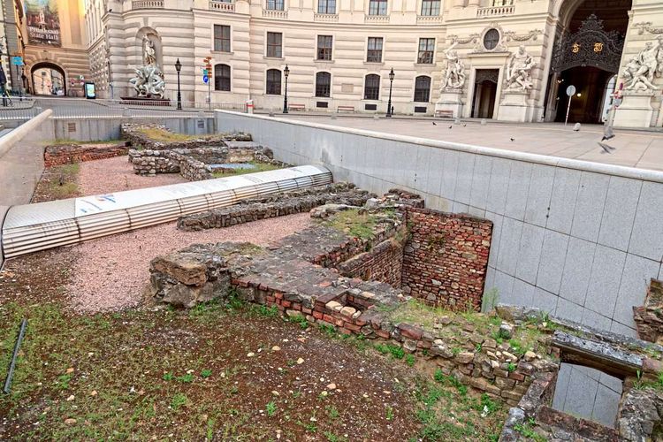 Reste römischer Mauern, eingefasst von einem modernen Bauwerk, im Hintergrund die Hofburgfassade