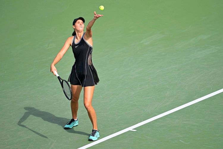 Eine Tennisspielerin in einem schwarzen Outfit führt während eines Spiels einen Aufschlag aus. Sie steht auf einem grünen Hartplatz.