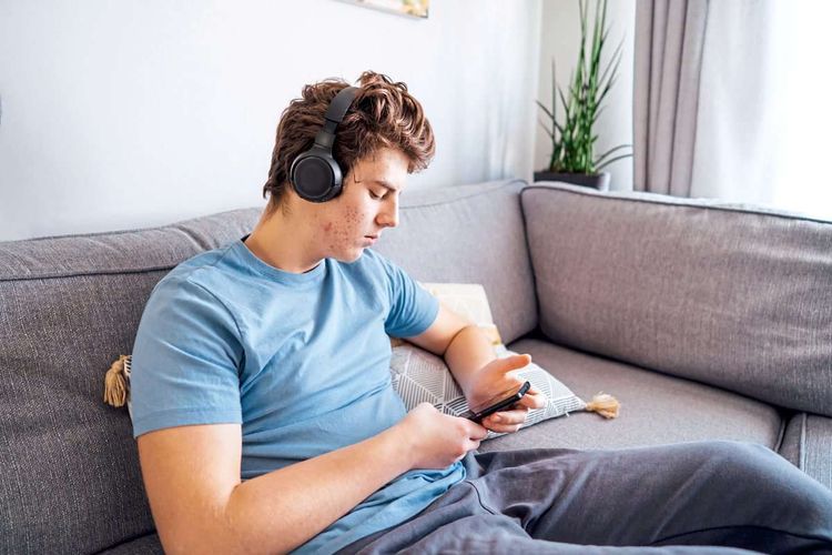 Teenager Junge mit unreiner Haut im Gesicht sitzt mit Kopfhörern auf dem Sofa und schaut auf sein Smartphone