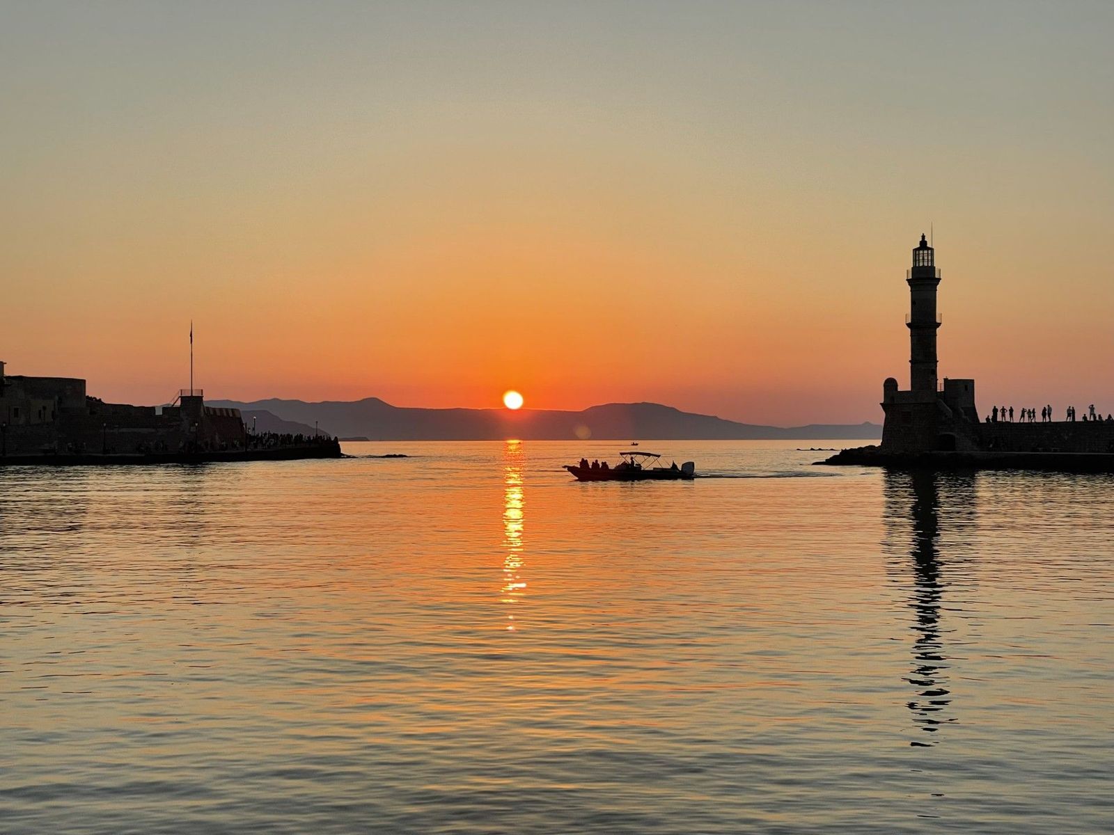 Sonnenuntergang am Meer, ein Boot fährt in einen Hafen ein, rechts steht ein Leuchtturm
