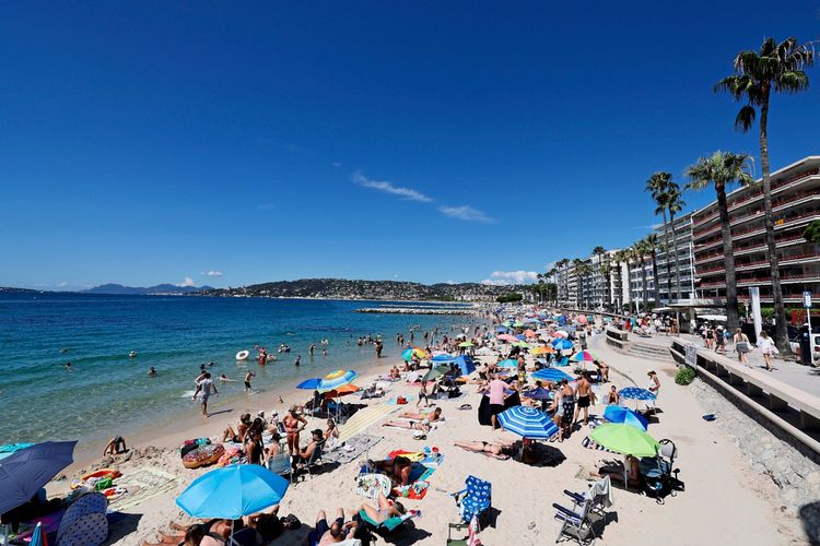 Menschen genießen die Sonne an einem belebten Strand in Cannes, Südfrankreich. Der Himmel ist blau, Palmen säumen die Strandpromenade, und bunte Sonnenschirme spenden Schatten.