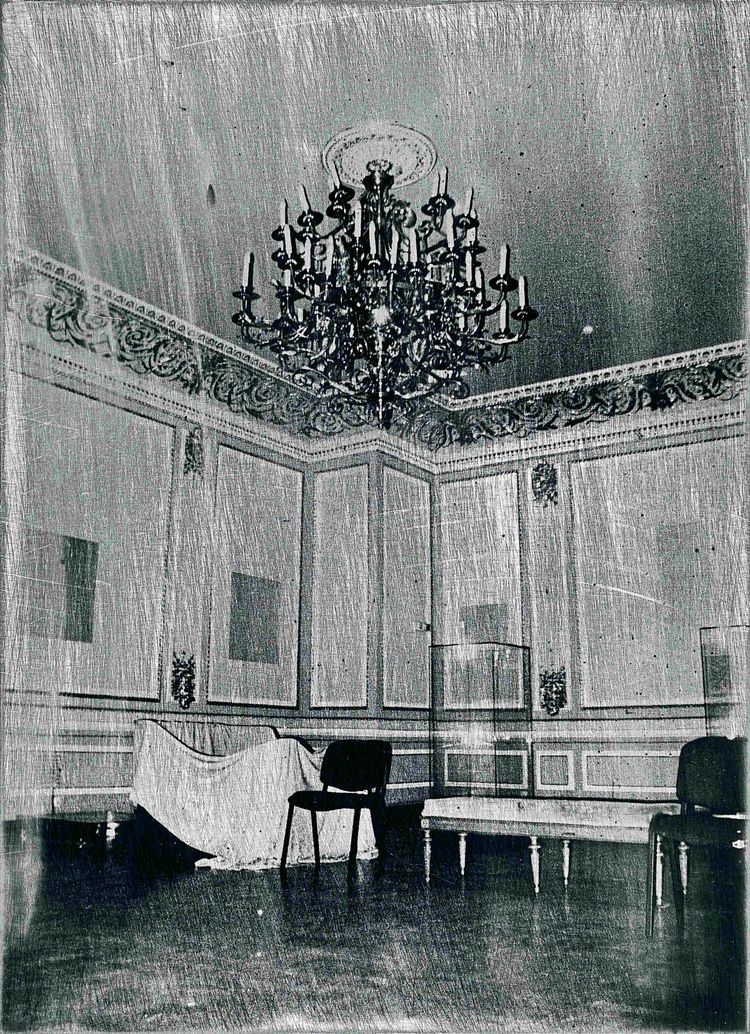 Schwarz-Weiß-Fotografie eines eleganten Raums mit aufwendig verziertem Kronleuchter, dekorativen Wandverkleidungen und Stuckarbeiten. Im Raum stehen ein paar einfache Stühle, eine überdeckte Struktur und eine leere Vitrine.