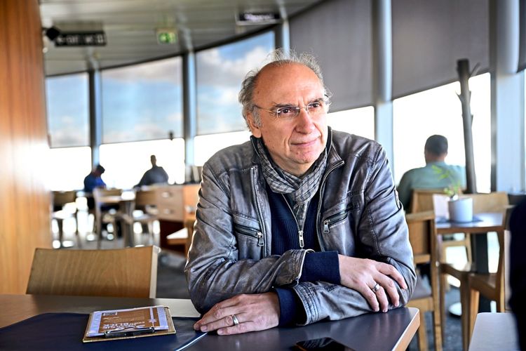Dimitré Dinev sitzt an einem Tisch im runden Café im Donauturm, im Hintergrund große Fenster. Er trägt eine schwarze Lederjacke, einen Schal und hat die Hände vor sich verschränkt. Auf dem Tisch liegt eine Speisekarte.