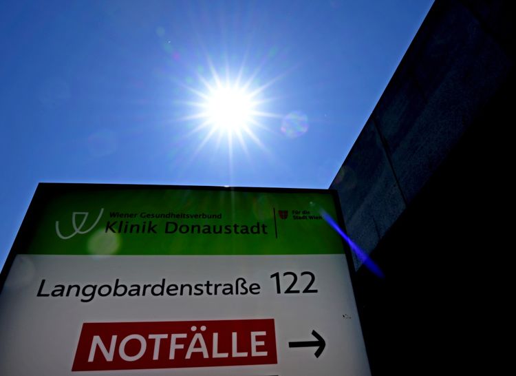 Bildbeschreibung: Die Sonne strahlt hell am klaren blauen Himmel hinter einem grünen und weißen Hinweisschild vor der Klinik Donaustadt in Wien. Auf dem Schild sind die Adresse 