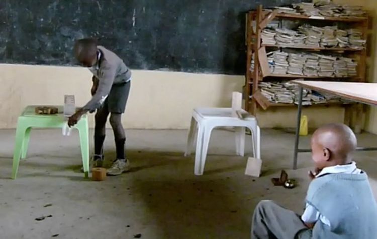 Zwei akrikanische Kinder in einem Klassenzimmer. Eines ist mit einem Murmelspiel beschäftigt, das andere schaut zu.