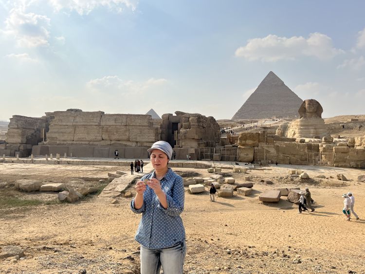 Die Forscherin trägt die Haare bedeckt und eine blau-weiß gepunktete Bluse, im Hintergrund sind die Pyramiden von Gizeh und die berühmte Sphinx zu sehen.