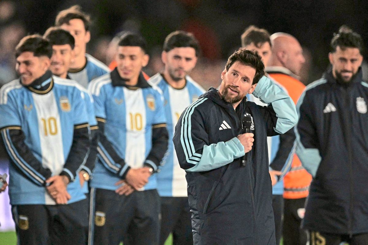 Eine Gruppe argentinischer Fußballspieler steht auf dem Spielfeld, einige tragen Trikots mit der Nummer 10. Ein Spieler vorne, der eine Trainingsjacke und ein Mikrofon hält, spricht und berührt seinen Kopf. Hintergrund unscharf.