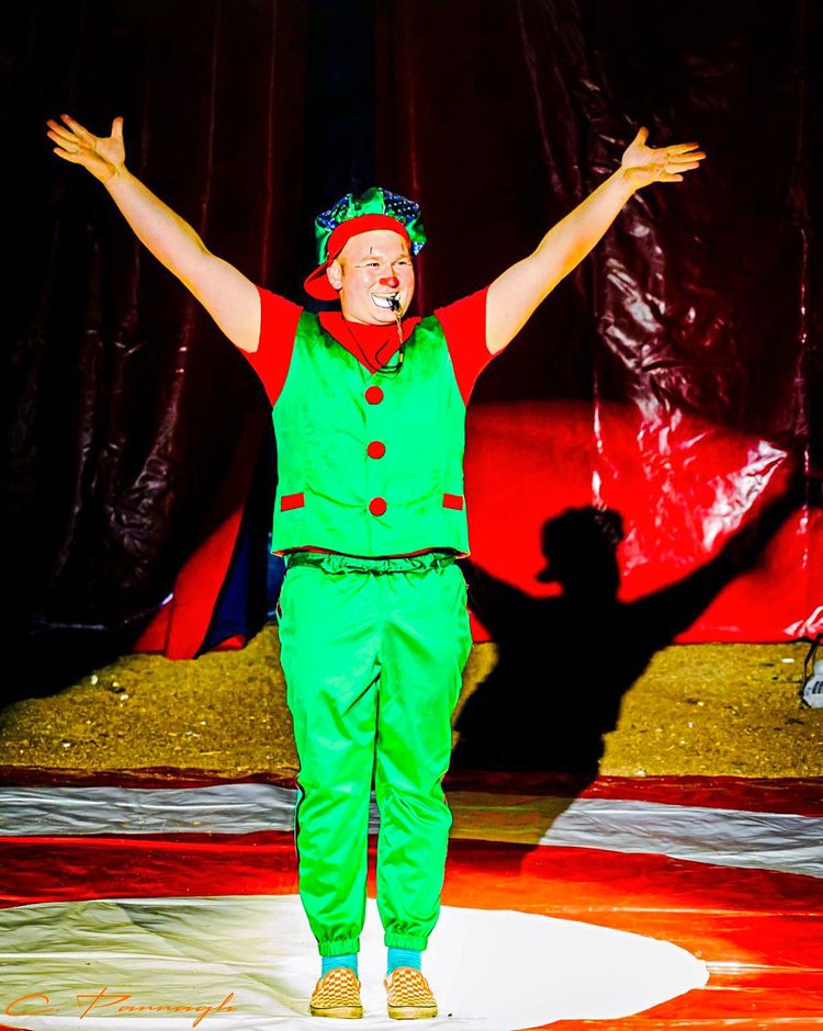 Marvin Spindler vom Zirkus Safari steht in der Manege als Clown verkleidet. In seinem grünen Gewand streckt er die Arme nach oben.