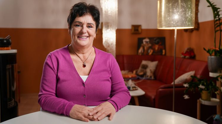 Die Frau im Bild trägt eine lila Strickweste, sitzt an ihrem Esstisch und blickt lächelnd in die Kamera.