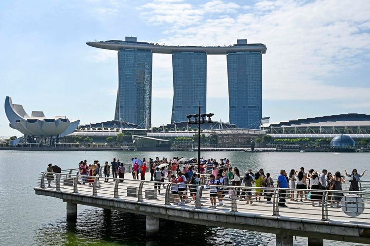 Menschen versammeln sich auf einem Steg an der Marina Bay Waterfront in Singapur und machen Fotos. Im Hintergrund sind das Marina Bay Sands Hotel und das ArtScience Museum zu sehen. Klarer Himmel und sonniges Wetter.