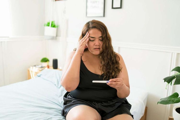 Übergewichtige Frau sitzt auf dem Bett und blickt besorgt auf einen positiven Schwangerschaftstest