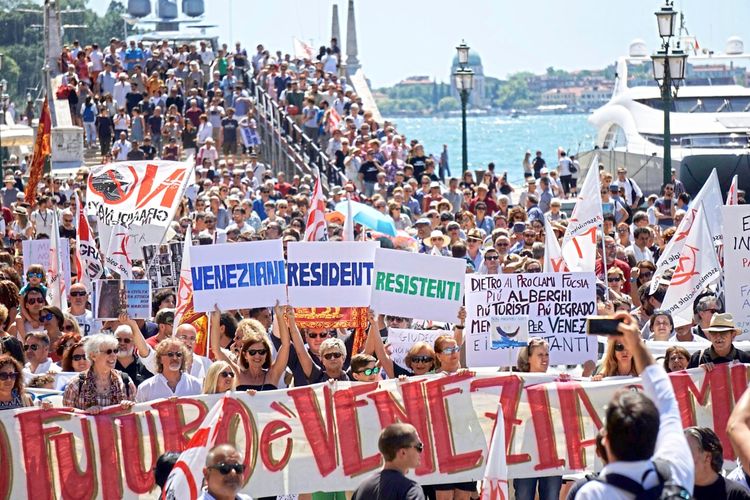 Menschenmenge bei einer Protestkundgebung in Venedig. Die Teilnehmenden halten Transparente und Schilder mit Aufschriften wie „Venetian Residents Resistenti“, um auf die Auswirkungen des Massentourismus aufmerksam zu machen. Im Hintergrund sind eine Brücke, ein Hafen und zahlreiche weitere Demonstrierende zu sehen.