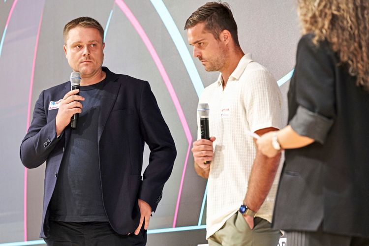 Zwei Männer sprechen bei einer Veranstaltung mit Mikrofonen. Links trägt eine Person einen dunklen Anzug, rechts eine Person ein helles Poloshirt. Im Hintergrund ist eine bunte Grafik zu sehen.