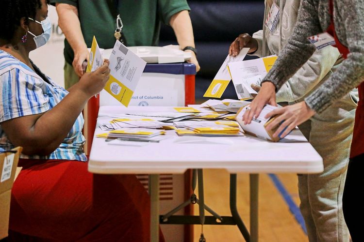 Menschen sitzen und stehen um einen Tisch in einem Wahllokal in Washington, D.C., und sortieren Briefwahlstimmen. Auf dem Tisch liegen zahlreiche Umschläge und Dokumente. Die Szene zeigt eine organisierte Auszählung am Abend der US-Präsidentschaftswahl am 6. November 2024.