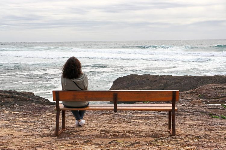 Frau sitzt alleine auf einer Bank mit Blick auf das Meer.