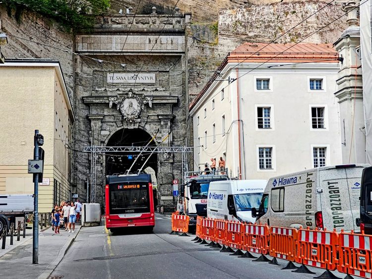 Eine Straße in Salzburg vor dem Neutor (Sigmundstor), mit einem roten Bus der Linie 10 im Vordergrund. Auf der rechten Seite sind Lieferwagen und orange-weiße Absperrgitter zu sehen, die auf eine Baustelle oder Veranstaltung hinweisen. Menschen stehen oder gehen entlang des Gehwegs. Historische Gebäude und der dahinterliegende Mönchsberg sind sichtbar.