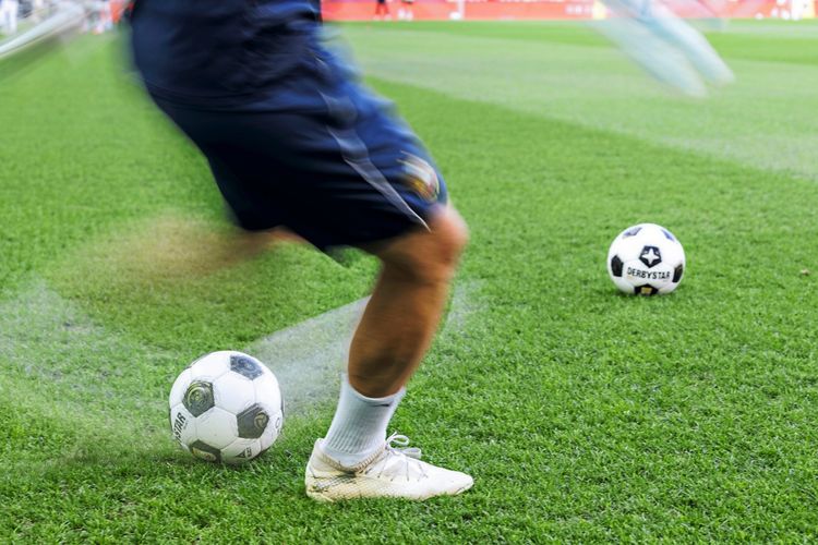 Ein Spieler in Bewegung auf einem Fußballfeld, zwei Fußbälle auf dem Rasen.