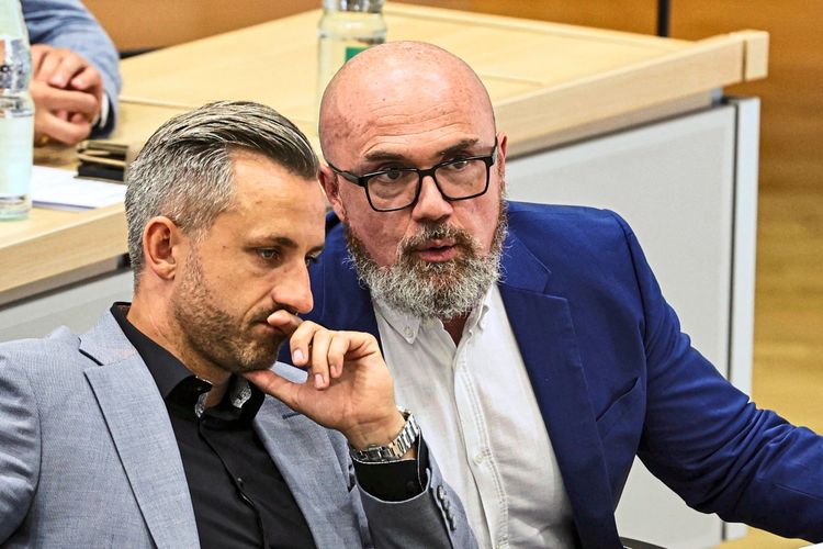 Zwei Männer in Anzügen sitzen nebeneinander während einer Landtagssitzung in Sachsen-Anhalt. Einer spricht, während der andere nachdenklich schaut. Auf dem Tisch vor ihnen liegen Unterlagen und eine Wasserflasche.