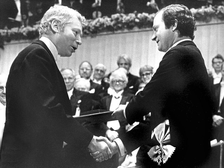 Schwarz-Weiß-Fotografie einer Preisverleihung: Ein Mann im Anzug schüttelt einem anderen die Hand und nimmt ein Dokument entgegen. Im Hintergrund sind weitere Personen in festlicher Kleidung sichtbar. Blumen schmücken den oberen Bereich des Raums, was auf eine formelle Zeremonie hindeutet.