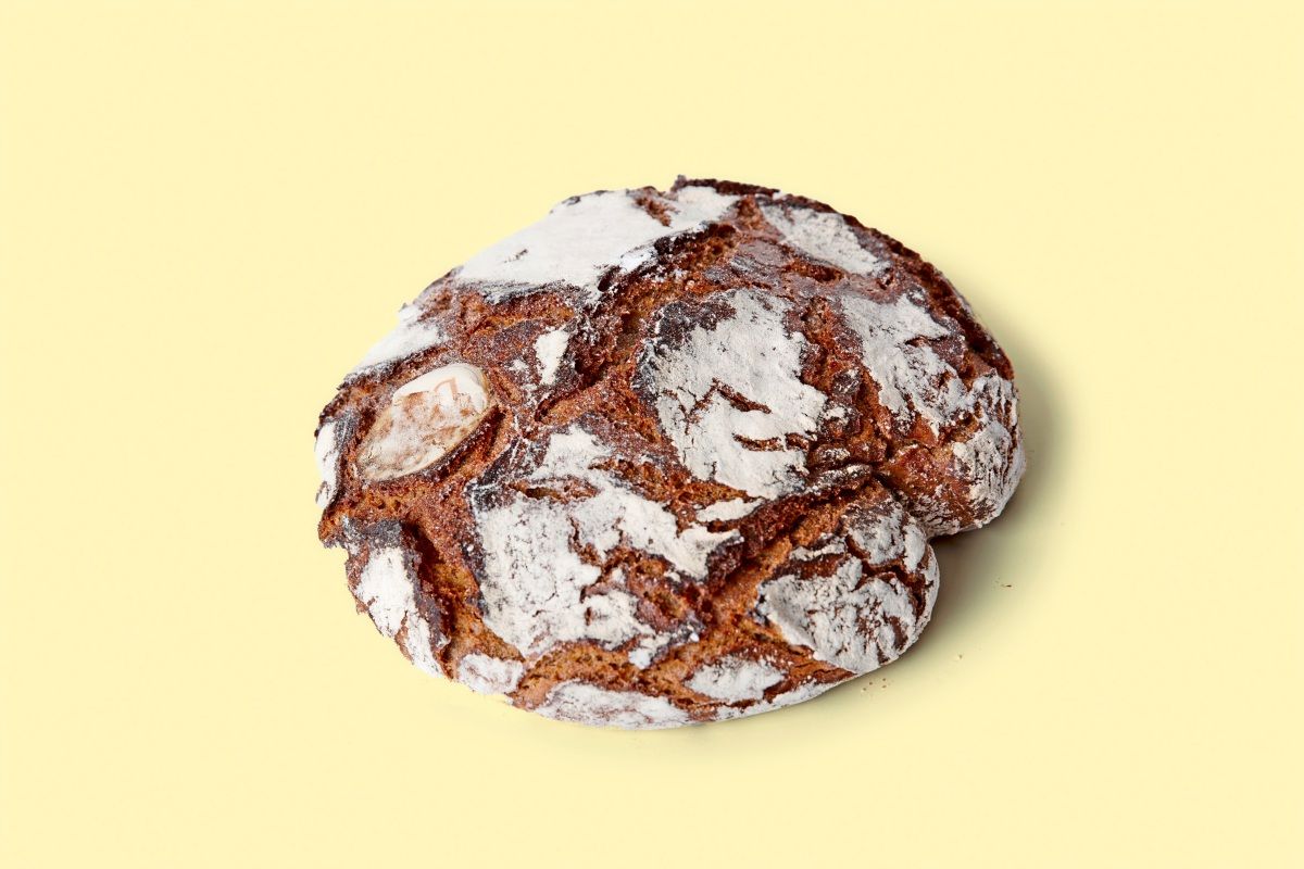 Ein rundes Roggenbrot mit knuspriger, dunkelbrauner Kruste und Mehlbestäubung, auf beigem Hintergrund.